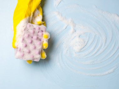 Sarı lastik eldivenli bir el mavi arka planda sabunlu köpüklü bir temizlik süngeri tutar. Ev temizliği konsepti, temizlik hizmeti