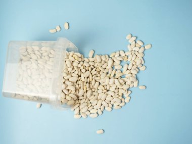 Mavi bir masanın üzerindeki plastik bir konteynırdan saçılmış kuru beyaz fasulyeler. Boşluğu kopyala