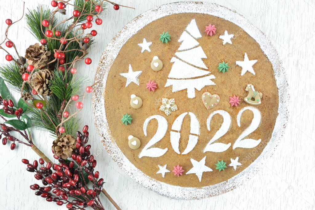 Traditional Greek new years cake, known as vasilopita, for 2022 with red tassels and artificial pine and red berries sticks on white wooden table