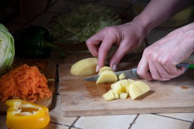 Bir kadın mutfakta taze sebzelerin arka planında patates kesiyor.