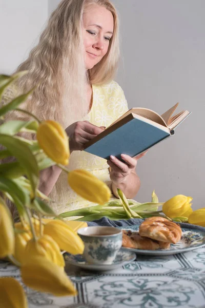 Uma mulher de meia-idade lê um livro em uma mesa com uma sobremesa e tulipas amarelas, — Fotografia de Stock