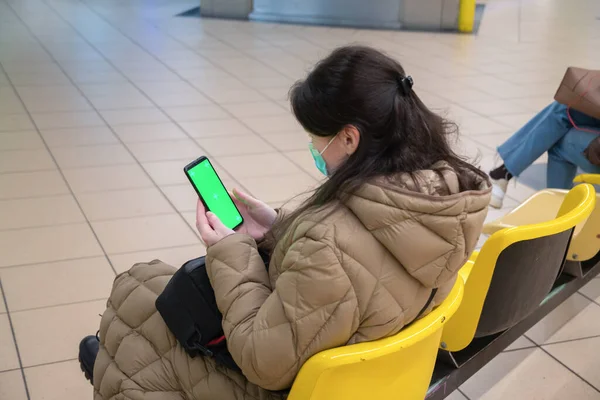 a young woman in a warm down jacket with green screen smartphone sits on an armchair in the waiting room in the subway watches the train and use social media, . High quality photo