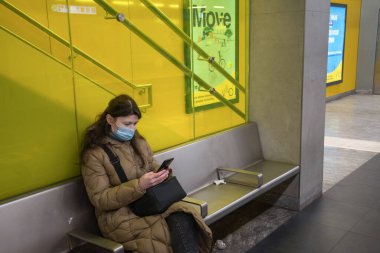 Ilık ceketli, yeşil ekran akıllı telefonlu genç bir kadın metroda bekleme odasında bir koltuğa oturmuş treni izliyor ve sosyal medyayı kullanıyor.