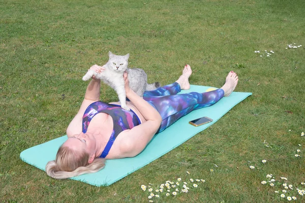 middle-aged woman doing yoga with a kitten on a blue mat in the middle of a green lawn in the fresh air, a person goes in for sports at any age. High quality photo