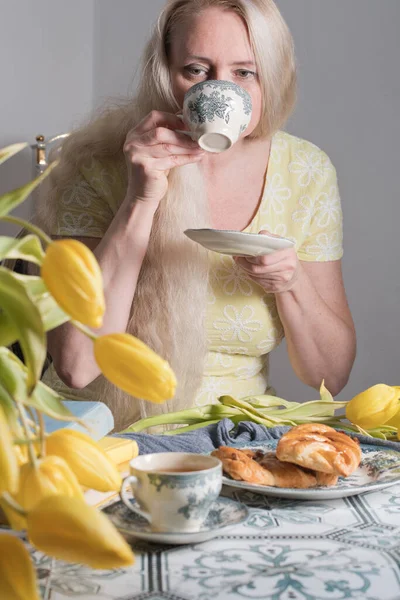Drinking coffee and tea, a middle-aged blonde woman with long hair drinks tea from a vintage cup and eats puff pastry with pecans on the background of yellow tulips and books on the table