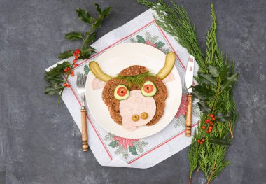 Sandviç, siyah ekmek, sosis ve sebzelerden yapılmış neşeli bir boğa portresi şeklinde, karanlık bir arka planda, thuja ve kutsal dallardan oluşan bir dekor ile.