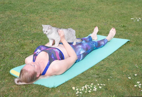 middle-aged woman doing yoga with a kitten on a blue mat in the middle of a green lawn in the fresh air, a person goes in for sports at any age. High quality photo