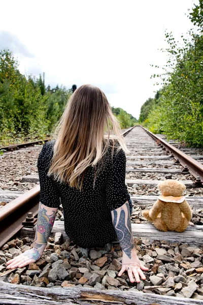 a young caucasian girl with glasses and tattoos smiles, a brown teddy bear sits in a copper pot against the background of an abandoned railway and old rails. High quality photo