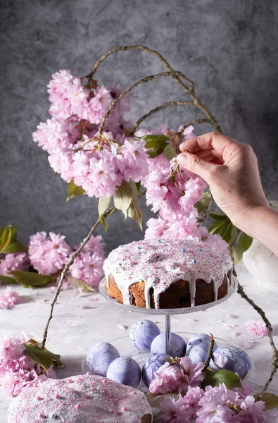 a woman decorates a homemade Easter cake with pink sakura flowers, spring blossom, a bouquet of pink sakura flowers on a table in a decorated spring room, a beautiful still life. High quality photo