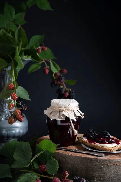 blackberry jam in a jar and a sandwich with butter on a saucer against the background of a branch with berries, food preparation for the winter period, a rich summer harvest. High quality photo