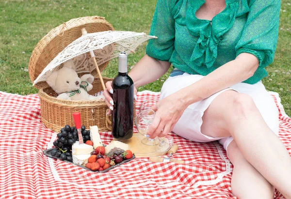 woman in a green blouse sits on a red checkered picnic rug, a cheese plate with red wine and berries, grapes, folded cherries, and an umbrella with a teddy bear in a straw basket on a summer sunny day