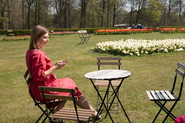 A young beautiful girl in a red dress in a spring park plays with her hair, flirts and takes a selfie on a mobile phone, tulips bloom and people walk in the park, pamper themselves.High quality photo