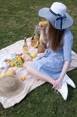 a young woman in a blue dress and straw hat is resting on a picnic with fruits, cheese plate and champagne, rest from worries and household chores parks and recreation areas,.High quality photo