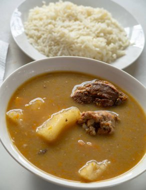 A white bowl of Dominican sancocho with yucca, chicken, pork, squash and more, served with white rice and a metal spoon.