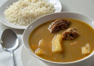 A white bowl of Dominican sancocho with yucca, chicken, pork, squash and more, served with white rice and a metal spoon.