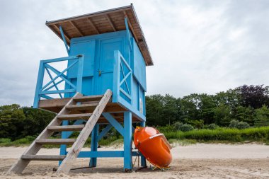 A blue lifeguard booth on the beach on a cloudy day. Orange lifeboat leaning against the side of the house.