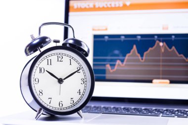 Dizüstü bilgisayarın arka planında hisse senedi grafiği olan çalar saat. Zaman, borsa, iş başarısı ve yatırım kavramı.