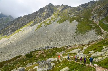 Bir grup turist bulutlu havada, kayalık dağların ve dik kayalık yamaçların arasında bir dağ yolu boyunca yürüyorlar. Yüksek Tatras, Slovakya