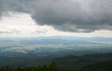 Bulutlu gökyüzünün altındaki dağ sırtından küçük kasabalar ve renkli tarlalarla vadiyi gör. Yüksek Tatras, Slovakya