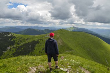 Bir dağın tepesinde dikilip yeşil dağ sırasına ve zirvelere bakan ve manzaranın tadını çıkaran bir adamın arka görüntüsü.