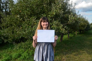 Gülümseyen, şık, esmer bir kızın elindeki beyaz kağıt parçasına bakıp doğal arka planda poz vermesi ön planda. Reklam kavramı.