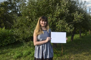 Gülümseyen, şık, esmer bir kızın elindeki beyaz kağıt parçasına bakıp doğal arka planda poz vermesi ön planda. Reklam kavramı.
