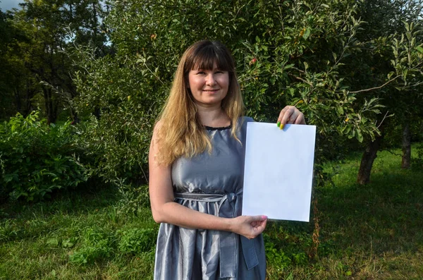 Gülümseyen, şık, esmer bir kızın elindeki beyaz kağıt parçasına bakıp doğal arka planda poz vermesi ön planda. Reklam kavramı.