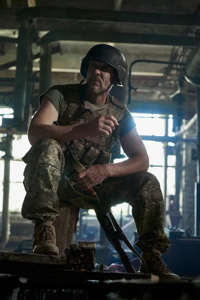 A Ukrainian soldier in a helmet and bulletproof vest smokes a cigarette in a house destroyed by a bomb.