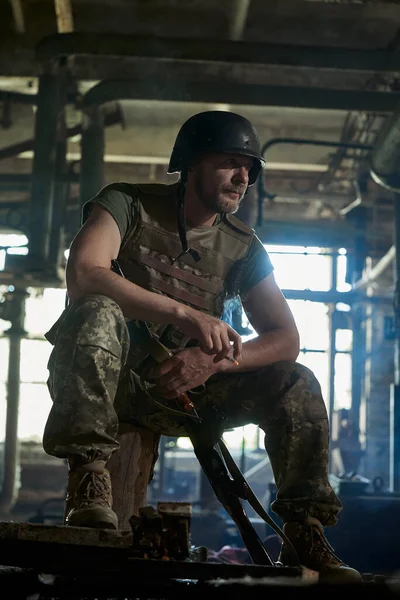 A Ukrainian soldier in a helmet and bulletproof vest smokes a cigarette in a house destroyed by a bomb.