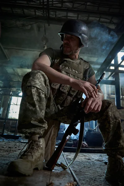 A Ukrainian soldier in a helmet and bulletproof vest smokes a cigarette in a house destroyed by a bomb.