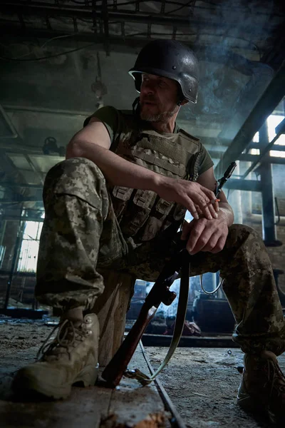 A Ukrainian soldier in a helmet and bulletproof vest smokes a cigarette in a house destroyed by a bomb.