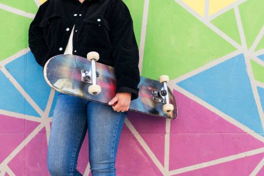 Renkli bir duvara yaslanmış kaykayıyla tanınmayan genç bir kadın.