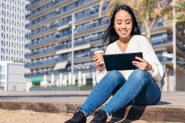 Genç Asyalı kadın gülümsüyor, elinde kahve tabletini seyrediyor.