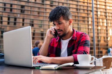 Genç latin adam telefonda konuşuyor ve bilgisayarını şehirdeki bir kahve dükkanının terasında daktilo ediyor. Teknoloji ve şehir yaşam tarzı kavramı.