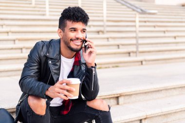 Mutlu genç Latin öğrenci merdivenlerde oturmuş gülümsüyor elinde kahve, teknoloji ve şehir yaşam tarzıyla telefonda konuşuyor.