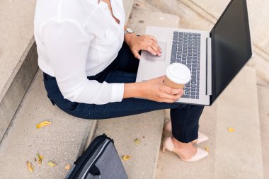 Laptop bilgisayarıyla çalışan tanınmayan bir iş kadını beton bir merdivende oturuyor. Bir elinde kahve, dijital girişimci konsepti ve kentsel yaşam tarzı.