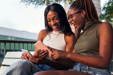 İki genç Afrikalı kadın cep telefonlarına bakarken mutlu bir şekilde gülüyor, gençlik kavramı ve iletişim, metin için kopyalama alanı