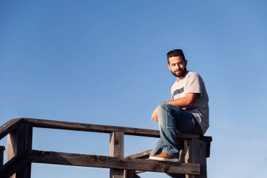 Kameraya bakan çekici bir genç arka planda mavi gökyüzü olan ahşap bir korkulukta oturuyor, boş zaman kavramı ve rahatlama, metin için kopyalama alanı