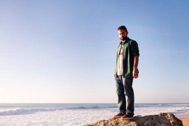 Yakışıklı sakallı adam ciddi bir şekilde kameraya bakıyor. Plaj kayası üzerinde duruyor. Boş zaman kavramı ve rahatlama.