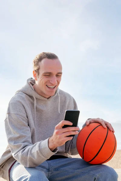 Young Caucasian Man Hoodie Basketball Smiling Using His Mobile Phone — стоковое фото