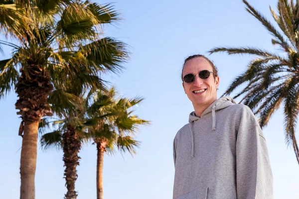 Güneş gözlüklü genç beyaz adam gülümsüyor. Palmiye ağaçlarının yanındaki kameraya bakıyor. Gençlik kavramı ve modern yaşam tarzı.