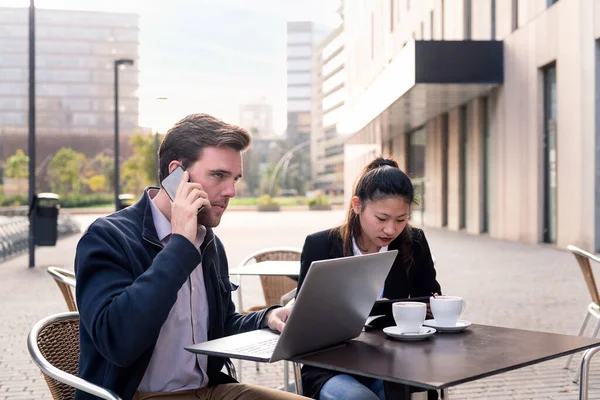 young businessman having a work meeting over coffee with his asiatic partner on a terrace in the financial district, concept of entrepreneurship and business