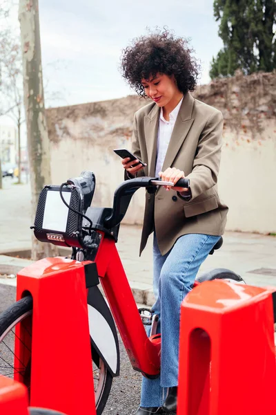 young latin woman checking her phone while sitting on a bike at the rental station, technology and sustainable mobility concept