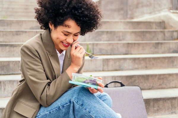 young woman laughing while eating a salad from a lunch box sitting on a staircase during a break from work, concept of urban lifestyle and healthy food, copy space for text
