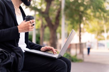unrecognizable stylish man working with a laptop computer drinking coffee outdoors in the city, concept of technology and urban lifestyle, copy space for text