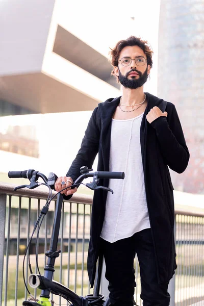young caucasian man with beard wearing trendy clothes and walking with his folding bike through the city, concept of urban lifestyle and sustainable mobility