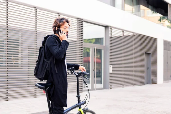 stylish young man standing with his folding bike talking on his mobile phone, concept of urban lifestyle and sustainable mobility, copy space for text