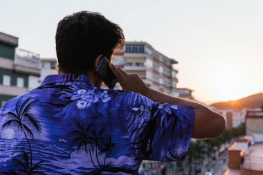 Sırtı mavi Hawai gömlekli, gün batımında telefonuyla konuşan tanınmamış genç bir adam. Konsept: tele-çalışma - kripto para birimi