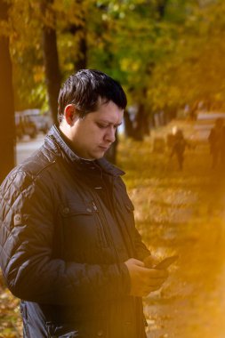 Sonbahar parkında yürüyen genç adam telefona, dikey fotoğrafa, portreye bakıyor..