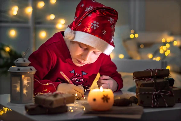 Chłopiec Czerwonym Świątecznym Kapeluszu Siedzi Przy Stole Pisze Listy Życzeń — Zdjęcie stockowe